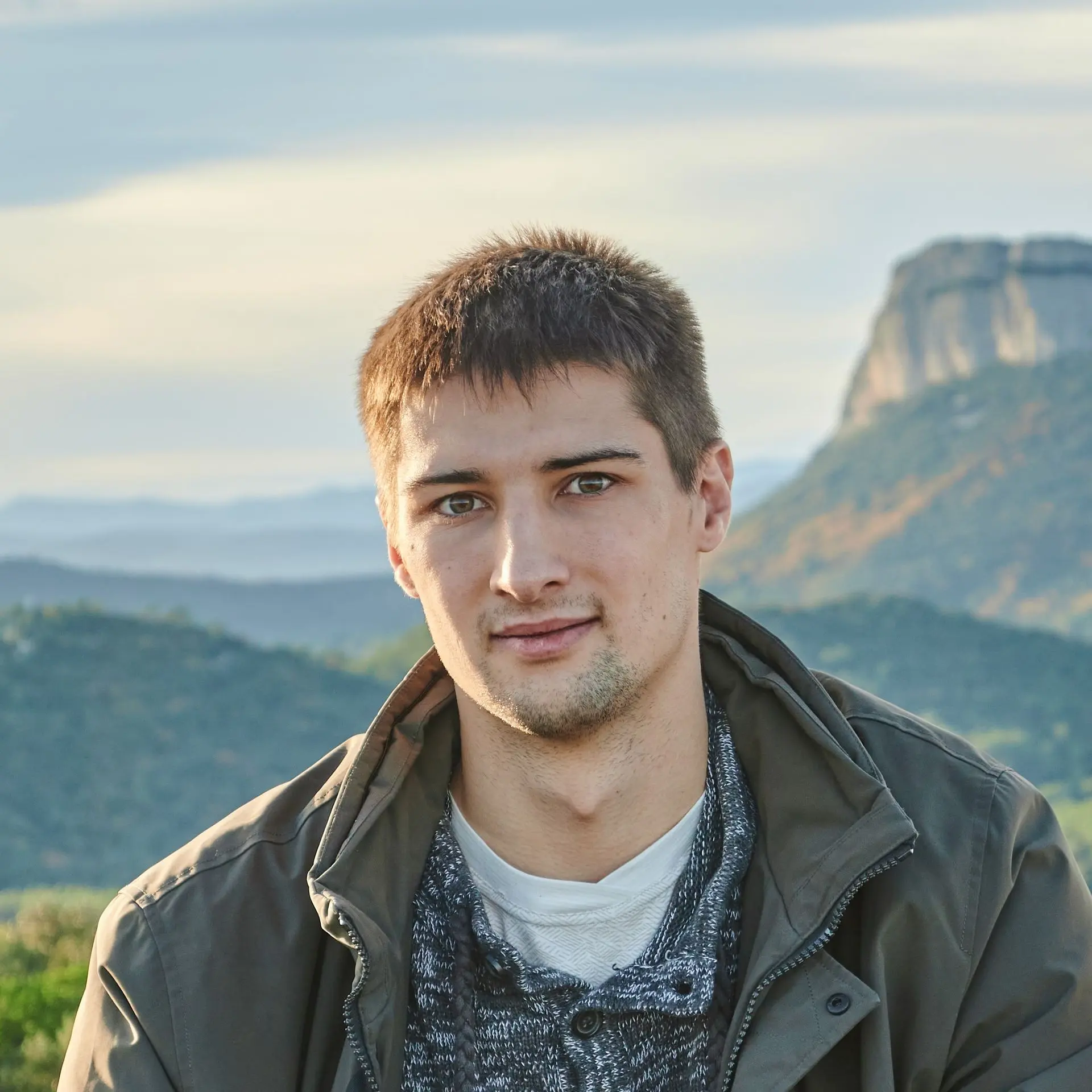 Portrait en format circulaire d'un jeune homme à l'extérieur. Il porte une veste sombre et se trouve devant un paysage montagneux.