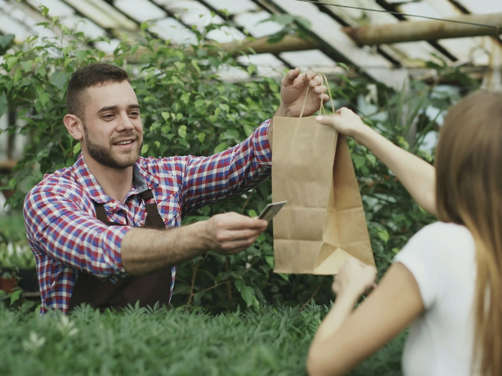 Un maraîcher remet un sac en papier à une cliente, dans sa serre.