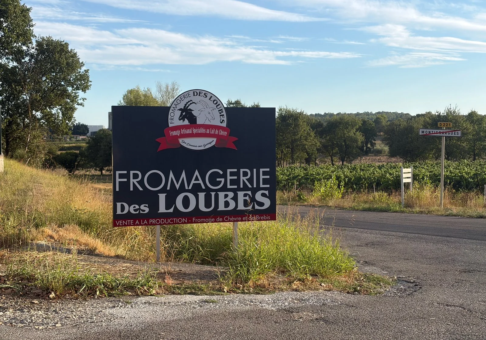 Le panneau de la Fromagerie des Loubes au bord d’une petite route bordée de vignes ; plus loin, le panneau de sortie de Montignargues.