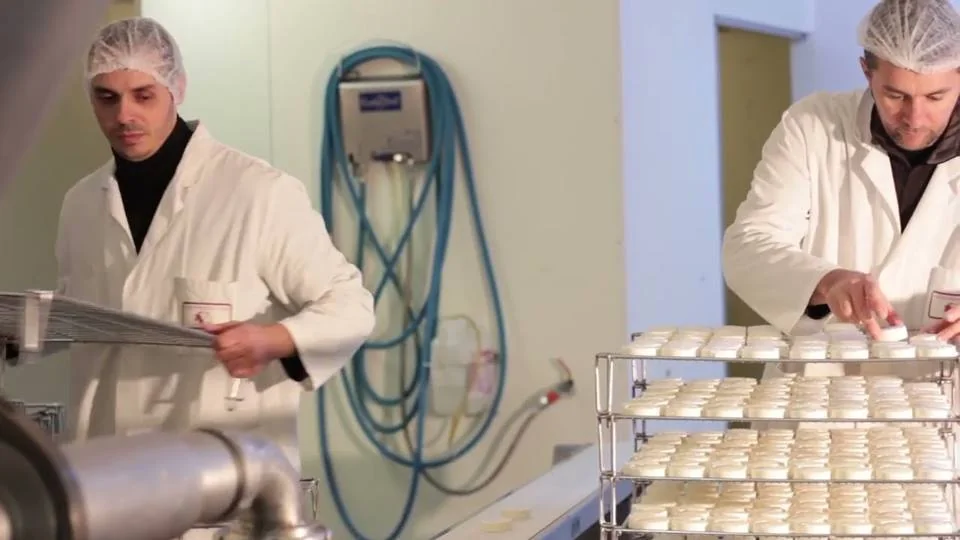 Dans l’atelier de la fromagerie, deux personnes en blouse blanche et charlotte : l’une tient une claie, l’autre pose des fromages sur des claies empilées.